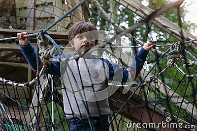 Low key light Portrait active kid climbing on rope frame at treehouse in the park. A boy standing alone on rope bridge in Stock Photo