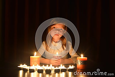 The cute little blonde girl holds a burning candle. Lots of candles are around her, over dark background Stock Photo