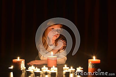 The cute little blonde girl keeps her hand on her shoulder and she is looking at the burning candle. Lots of candles are around h Stock Photo