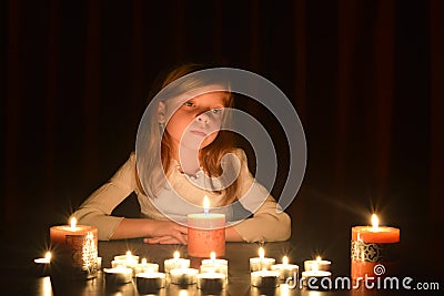 The cute little blonde girl is looking on the light of candle. Lots of candles are around her, over dark background Stock Photo