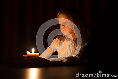 The cute little blonde girl is looking on the light of candle over dark background Stock Photo