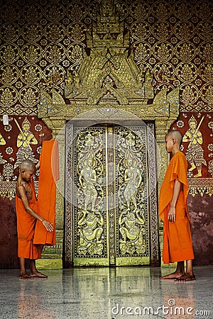 young buddhist monks folding an orange textile in front of a nicely decorated golden plated wooden door Editorial Stock Photo