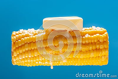 A lump of butter is melted on hot cooked corn. A large ear of yellow corn is shown against a light blue background Stock Photo
