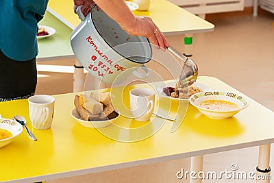 Lunch in a Russian kindergarten. The nurse lays out the food with a ladle from a saucepan with the inscription in Russian Stock Photo