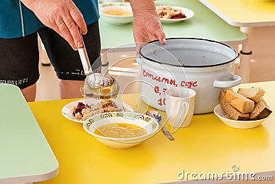 Lunch in a Russian kindergarten. The nurse lays out the food with a ladle from a saucepan with the inscription in Russian Stock Photo