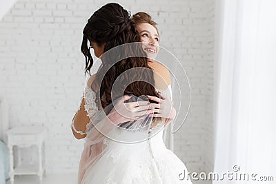 Luxury bride hugging bridesmaid and smiling, joyful moment in minimalistic loft white brick background Stock Photo