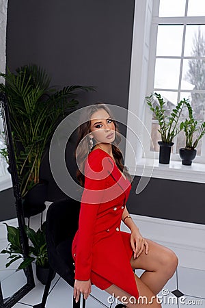 Luxury brunette with bright makeup posing in red dress in interior near window Stock Photo