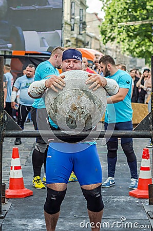 LVIV, UKRAINE - JUNE 2016: Strong bodybuilder strongman lifts a huge heavy stone ball made of marble and throws it over the bar on Editorial Stock Photo