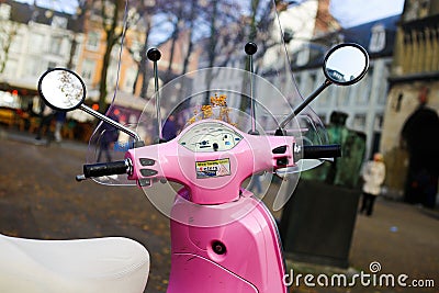 View beyond handlebar, tachometer, windshield and rearview mirrors of retro pink Vespa scooter on blurred city square Editorial Stock Photo