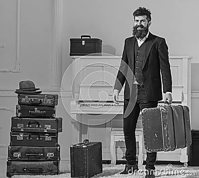 Macho elegant on smiling face stands near pile of vintage suitcase, holds suitcase. Man, traveller with beard and Stock Photo