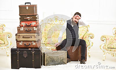 Macho elegant on strict face sits tired near pile of vintage suitcase. Man, butler with beard and mustache delivers Stock Photo