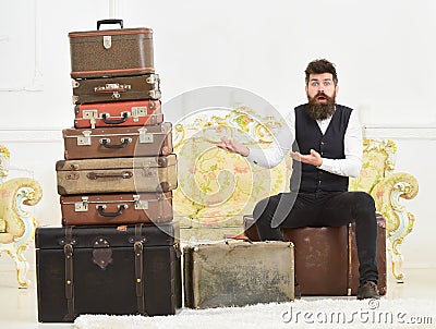 Macho elegant on surprised face sits, confused at end of packing, near pile of vintage suitcases. Man with beard and Stock Photo