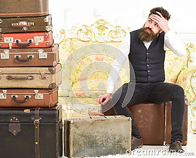 Macho elegant on tired face sits, exhausted at end of packing, near pile of vintage suitcases. Man with beard and Stock Photo
