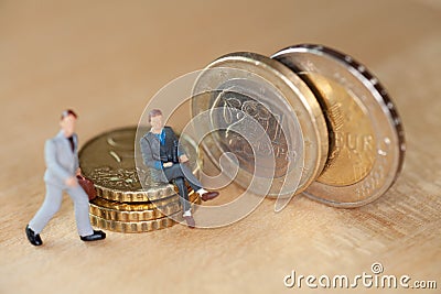 Macro of small figures sitting on a stack of coins next to it are two two euro coins with a symbol for Greece Stock Photo