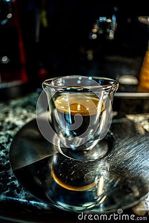 Macro view of a modern designed glass cup with freshly brewed black coffee with golden foam with small bubbles perched on a metal Stock Photo
