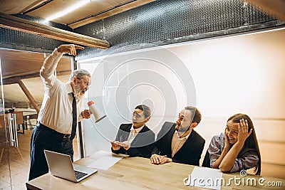 Angry boss with megaphone screaming at employees in office, scared and annoyed colleagues listening at the table Stock Photo