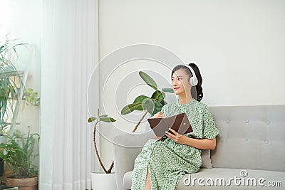 Mage of focused asian woman using headphones and writing down notes while sitting on sofa at home Stock Photo