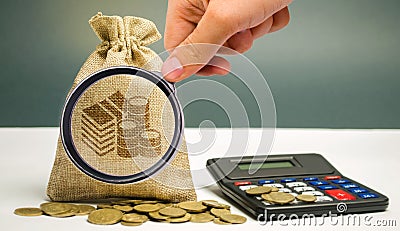 Magnifying glass looks at money bag with coins and a calculator. Profit calculation and income analysis. Interest rates. Stock Photo