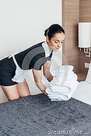 Maid in white apron putting pile of folded towels on bed in hotel room Stock Photo