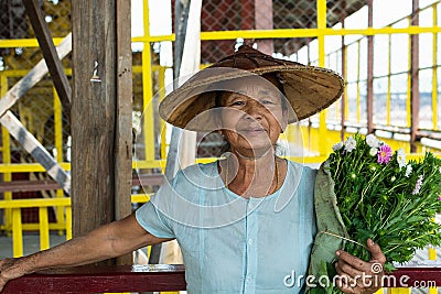 Maing Thauk, Myanmar - April 2019: portrait of an old Burmese woman in a bamboo hat and flowers Editorial Stock Photo
