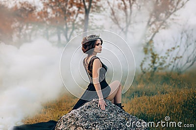 The majestic lady, the dark queen, sits on the stone baring her leg. The brunette girl in the gothic crown. The Stock Photo