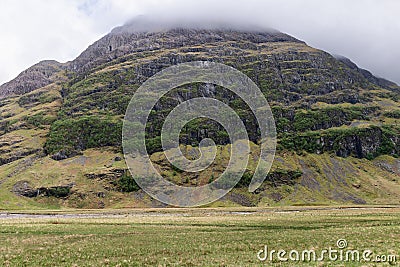 From Glen Coe valleys, a majestic mountain rises, draped in green with a cloud-capped summit Stock Photo