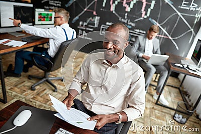 Make the smart move. Cheerful african male trader sitting by desk and studying analytical reports by looking through Stock Photo