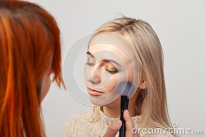 Makeup artist girl with red hair puts make-up on a blonde model with eyes closed, holds a brush in her hands and puts contouring Stock Photo