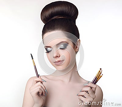 Makeup artist. Pretty teen girl with cute bun hairstyle and fashion beauty makeup, brunette holding brushes in hand isolated on w Stock Photo
