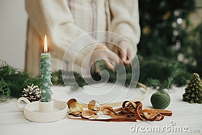 Making Christmas modern wreath. Stylish candle as fir tree, golden bells and ribbon on background of woman hands making boho Stock Photo