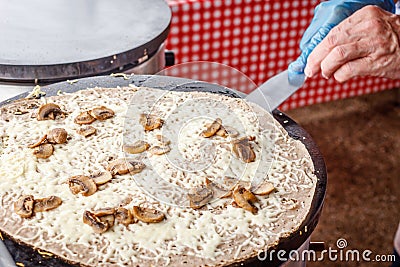Making crepes pancakes at a food market. A hand is making crepes outdoors with melted cheese and mushrooms on a metal griddle Stock Photo