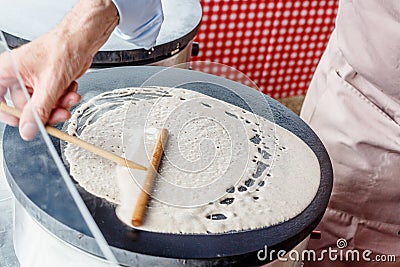 Making crepes pancakes in food market. A hand is making crepes outdoors spreading the batter onto a metal griddle with a wooden Stock Photo
