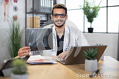 Male Asian Indian doctor, sitting in office, holding skull xray image, working on laptop Stock Photo