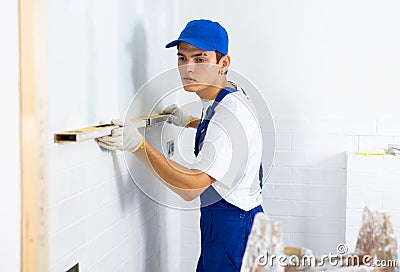 Male builder in blue overalls measures deviations from a given vertical wall using building level Stock Photo