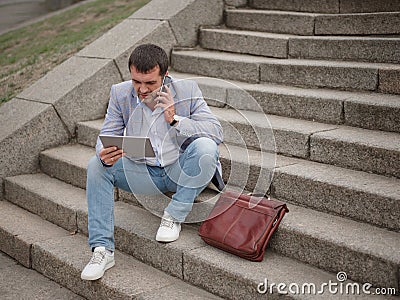 Employee with tablet and talking on a phone, sitting on the urban background. Technology progress concept. Copy space. Stock Photo