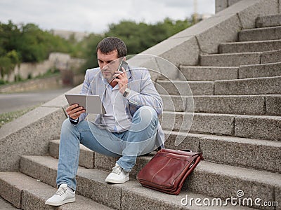 Employee with tablet and talking on a phone, sitting on the urban background. Technology progress concept. Copy space. Stock Photo