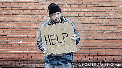 Homeless holds piece of cardboard with inscription Help. Refugee is at brick wall looking for job. Male tramp in dirty clothes, Stock Photo