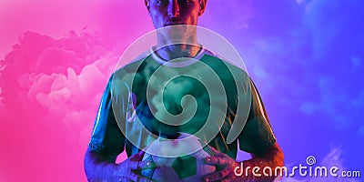 Male caucasian soccer player holding ball standing againsy smoky pink and blue background Stock Photo