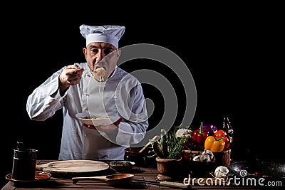 Male chef in a restaurant kitchen wearing white uniform tasting spaghetti with fresh vegetables Stock Photo