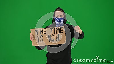 A male demonstrator with his face covered by a scarf holds up a sign that says Time is now. The man on a green screen Stock Photo