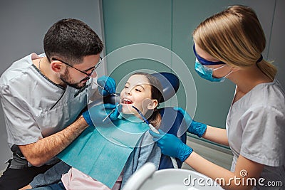 Male dentist check up girl`s teeth with dentist`s tools. Female helper stand beside. Girl sit in dental chair in room Stock Photo