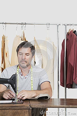 Male dressmaker stitching cloth on sewing machine with clothes rack in background Stock Photo