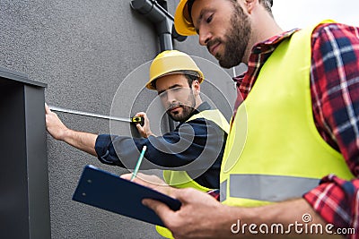 male engineers in safety vests and helmets working with measuring tape and clipboard Stock Photo