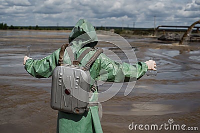 A male environmentalist in a green protective suit and gas mask takes a sample of water in a polluted lake. Waste from Stock Photo