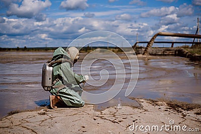 A male environmentalist in a green protective suit and gas mask takes a sample of water. The scientist is doing a Stock Photo