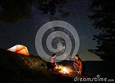 Male and female warming themselves around the fire at night camping above starry sky Stock Photo