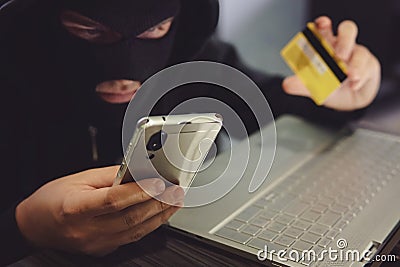 Male hacker in a robber mask uses phone, credit card and laptop in some fraudulent scheme. Cyber thief stole the personal data and Stock Photo