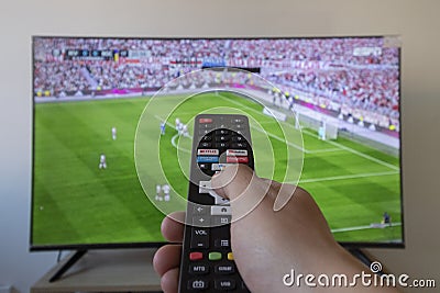 a male hand holding a black tv remote control with football game at background Editorial Stock Photo