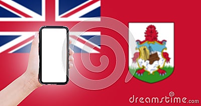 Male hand holding smartphone with blank on screen, on background of blurred flag of Bermuda. Close-up view. Stock Photo