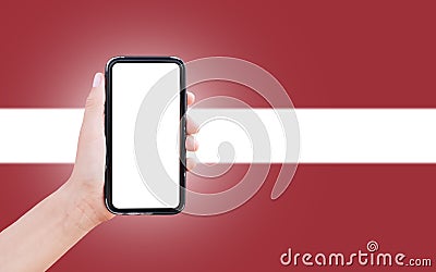 Male hand holding smartphone with blank on screen, on background of blurred flag of Latvia. Close-up view. Stock Photo